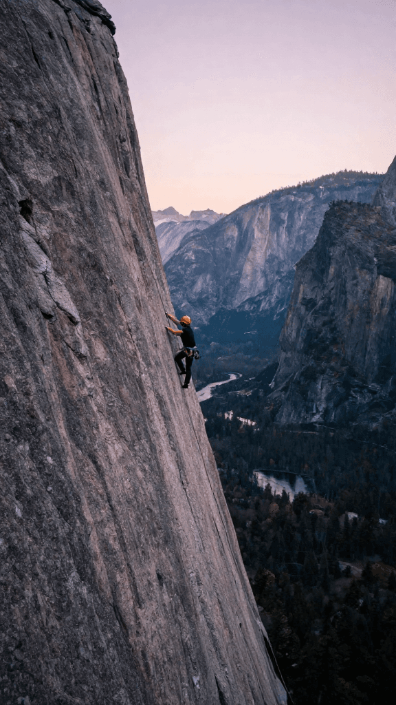 Climber ascends a sheer granite face in Yosemite early morning, the canyon breathing with pale pink light and distant water. Framing from a