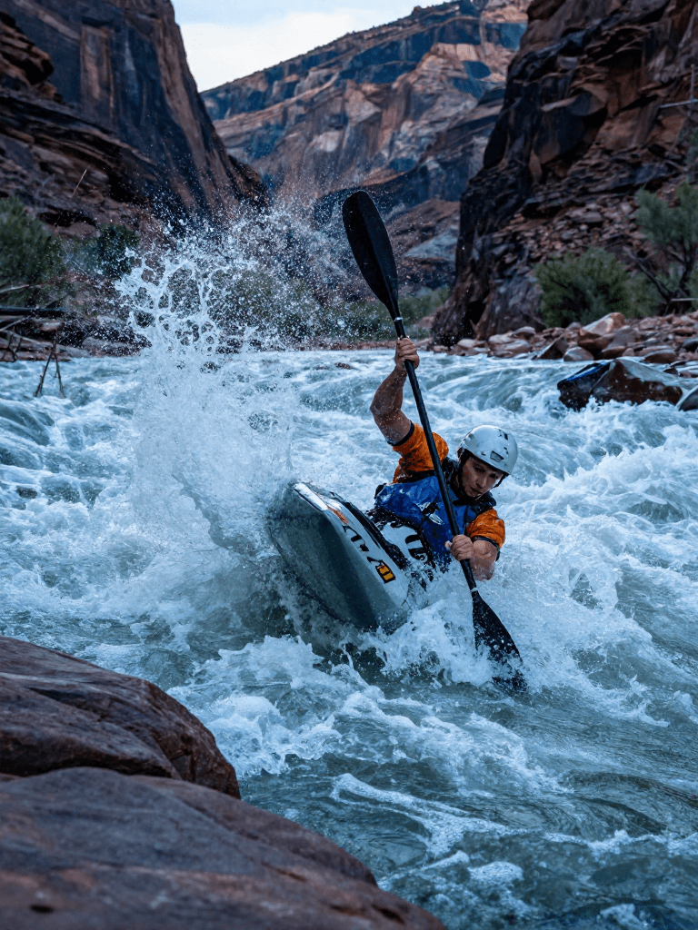 Whitewater carving on a raging Colorado River rapid, a kayaker half-hangs from the stern and powers through a foaming curtain. A riverbank v
